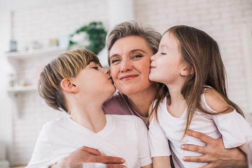 We love our granny. Portrait of happy gray-haired woman looking at camera and hugging her grandchildren. Grandchildren kissing her in house. Visiting grandmother concept