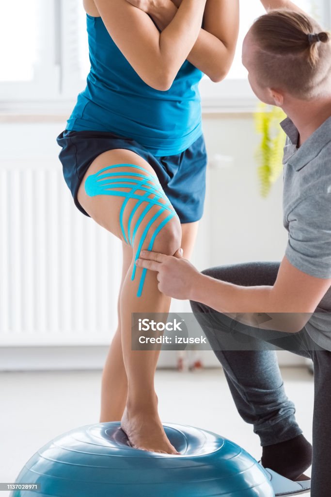 Young woman excercising with physiotherapist Physical therapist showing young woman an exercises on fitness ball. Patient having elastic therapeutic tape on her knee. Physical Therapy Stock Photo