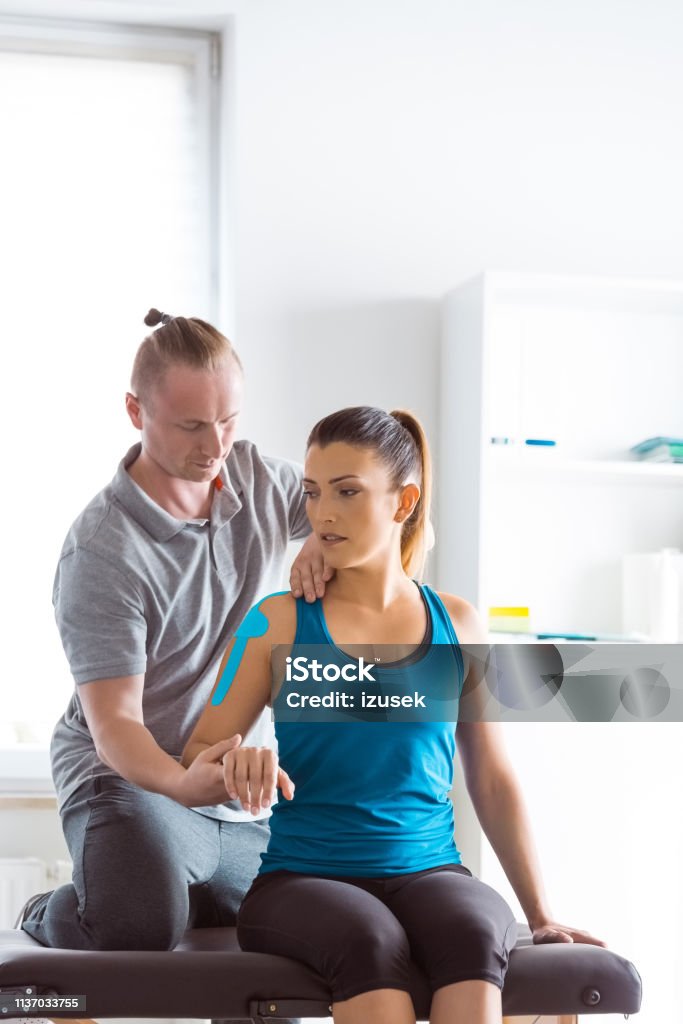 Physiotherapist massaging young woman Physical therapist giving shoulder massage to young woman. Patient having elastic therapeutic  tape on her arm. Physical Therapy Stock Photo