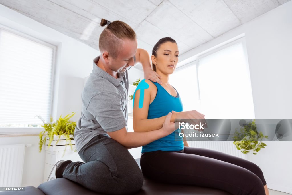 Physiotherapist massaging young woman Physical therapist giving shoulder massage to young woman. Patient having elastic therapeutic  tape on her arm. Medical Exam Stock Photo
