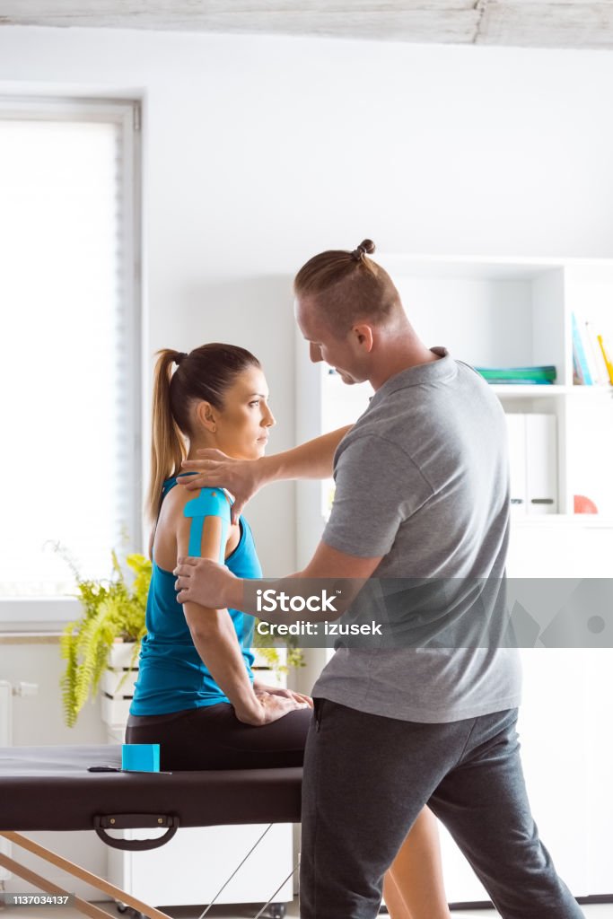 Physiotherapist massaging young woman Physical therapist giving shoulder massage to young woman. Patient having elastic therapeutic  tape on her arm. Alternative Therapy Stock Photo