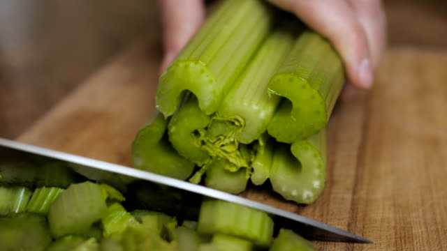 Knife chopping celery in restaurant kitchen on wooden cutting surface. Chef cutting celery to cook in the kitchen. Hands Slicing celery with Knife on chopping wooden board background. Slow motion.