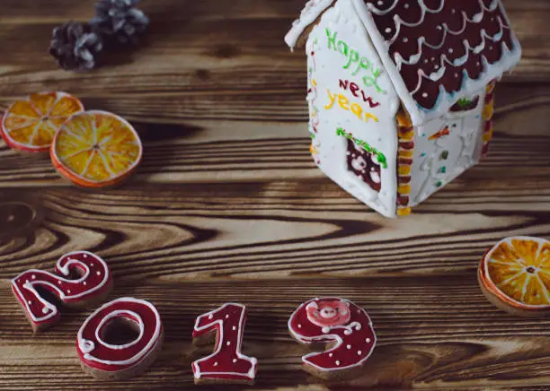 Christmas card on wood background gingerbread red numbers 2019 with slices of orange and white gingerbread house with a brown roof, a window and the inscription happy New Year on a white wall