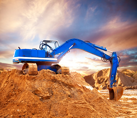 Large blue excavator caterpillar on a construction site. The excavator digs sand against the background of the setting sun. Orange - blue composition.