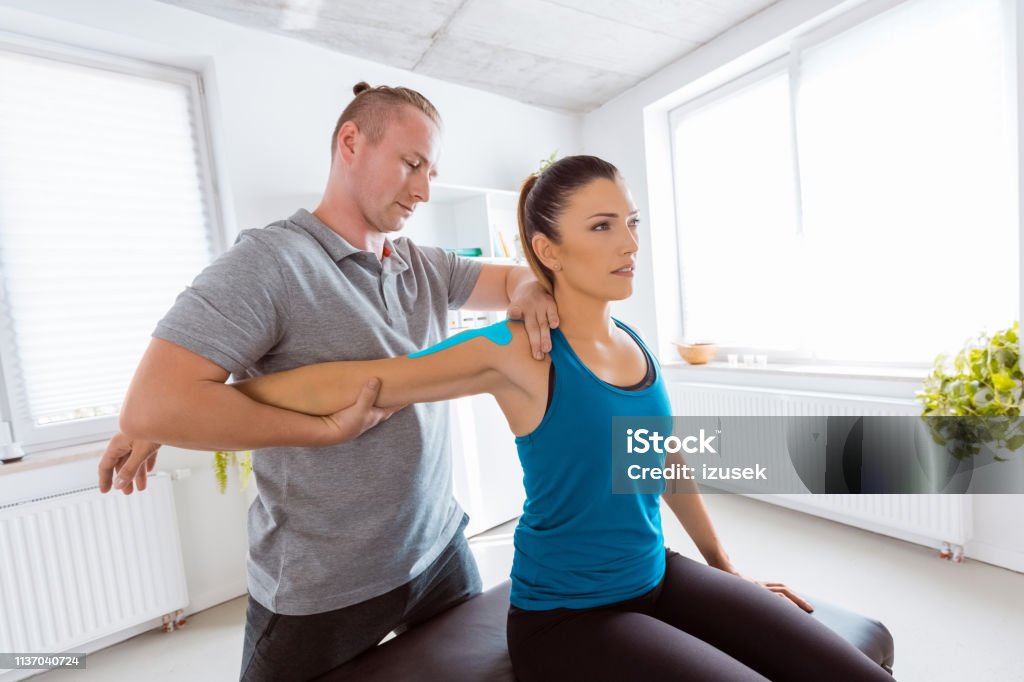 Physiotherapist massaging young woman Physical therapist giving shoulder massage to young woman. Patient having elastic therapeutic  tape on her arm. Elastic Therapeutic Tape Stock Photo