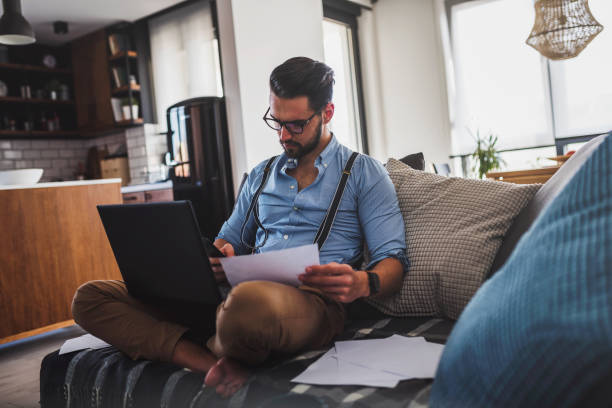 istock Modern bearded young businessman working on laptop computer while sitting on sofa at home, looking at some documents 1137040870