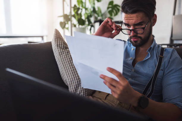 istock Modern bearded young businessman working on laptop computer while sitting on sofa at home, looking at some documents 1137042024