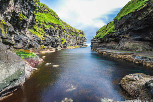 splendida vista sul molo o sul porto con roccia nera naturale ed erba verde nel villaggio di gjogv con acqua limpida in profondità nell'oceano atlantico settentrionale blu, cielo nuvoloso, isole faroe, destinazione di viaggio nascosta - clear sky panoramic grass scenics foto e immagini stock