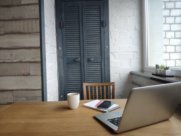 Close up of wooden desk built into recess of white wall in front of window. Nearby red chair, internet router and decor elements on table. On the background, outside window city buildings.