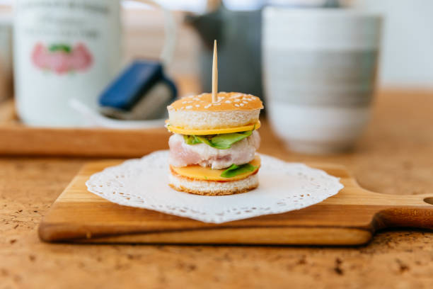 Mini Chicken Burger on wooden chopping board with blur tea cup and mug in the background.