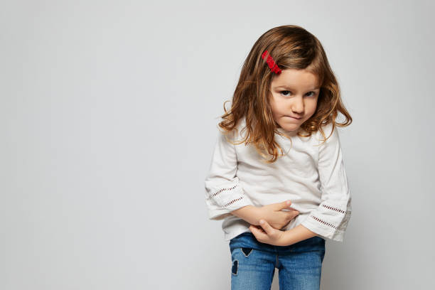 Young child standing against a white background looking at camera with her hands gripping her stomach