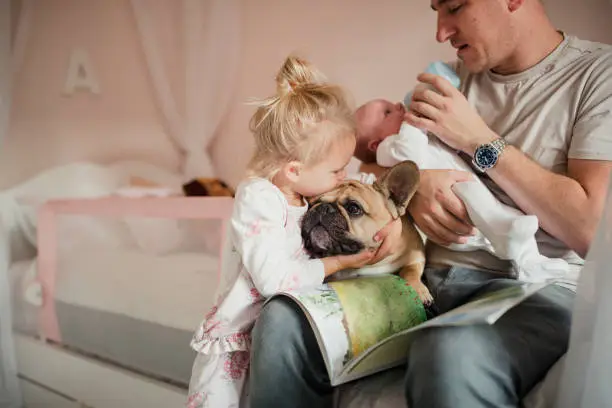Father feeding his baby while his young daughter and puppy cuddle and kiss each other beside him.