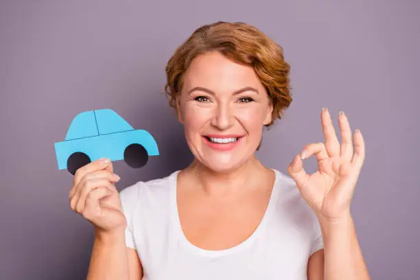 Close-up portrait of nice lovely attractive cheerful cheery wavy-haired lady in casual white t-shirt holding in hand blue car figure showing ok-sign isolated on gray violet purple pastel background.
