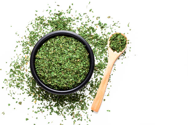 Spices: Top view of a black bowl filled with dried parsley isolated on white background. A wooden spoon is beside the bowl and dried parsley is scattered on the table. Predominant colors are white and green. High key DSRL studio photo taken with Canon EOS 5D Mk II and Canon EF 100mm f/2.8L Macro IS USM.