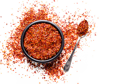Spices: Top view of a black bowl filled with red chili pepper flakes isolated on white background. A vintage metal spoon with pepper flakes is beside the bowl, pepper flakes are scattered on the table. Predominant colors are white and red. High key DSRL studio photo taken with Canon EOS 5D Mk II and Canon EF 100mm f/2.8L Macro IS USM.