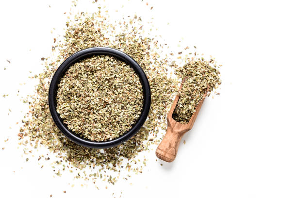 Spices: Top view of a black bowl filled with dried oregano isolated on white background. A wooden serving scoop is beside the bowl and dried oregano is scattered on the table. Useful copy space available for text and/or logo. Predominant colors are white and green. High key DSRL studio photo taken with Canon EOS 5D Mk II and Canon EF 100mm f/2.8L Macro IS USM.