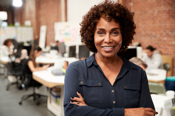 portrait of mature businesswoman in open plan office with business team working in background - business women manager looking at camera imagens e fotografias de stock