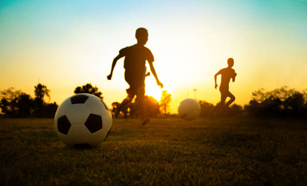 An action sport picture of a group of kids playing soccer football for exercise in community rural area under the sunset. Silhouette action sport outdoors of a group of kids having fun playing soccer football on green grass field kicking photos stock pictures, royalty-free photos & images