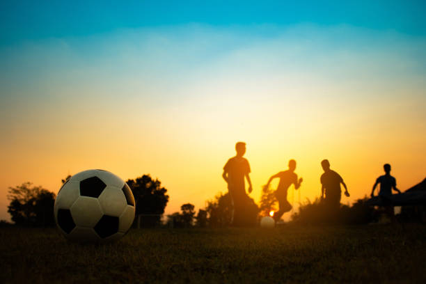una imagen deportiva de acción de un grupo de niños jugando al fútbol para el ejercicio en el área rural de la comunidad bajo la puesta del sol. - ball horizontal outdoors childhood fotografías e imágenes de stock