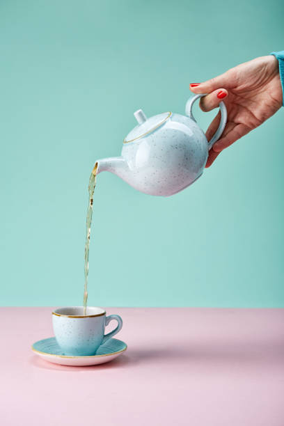 hand holding tea pot Tea pot pouring tea in a cup on blue backdrop pink tea stock pictures, royalty-free photos & images