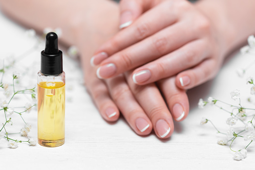 Yellow cuticle oil bottle and a female hands on a white wooden table background. Fingernail care concept.
