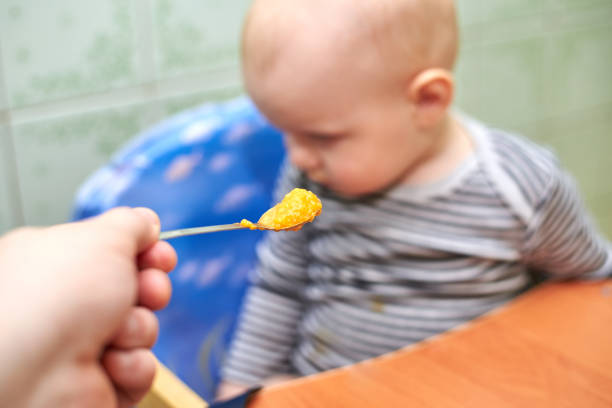 Portrait of little baby boy eating food. Baby with a spoon in feeding chair. Cute baby eating first meal.