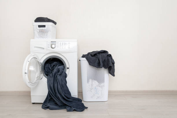 White washing machine standing against wall inside bright apartment light interior near laundry basket with black linen and copy space for text