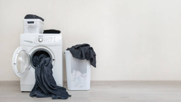 istock Concept of preparation to laundry process. White washing machine standing against wall inside apartment with light interior near plastic basket with black clothes 1137140034