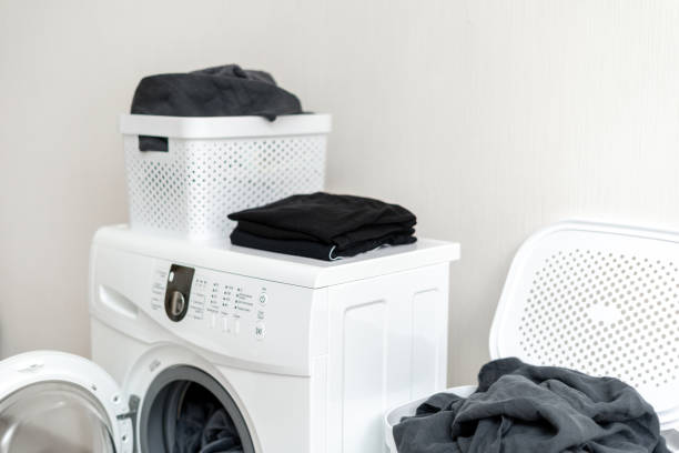istock Cropped and side view photo of preparation for laundry process. Modern washing machine standing inside bright apartment light interior against wall near plastic basket of black linen 1137140151