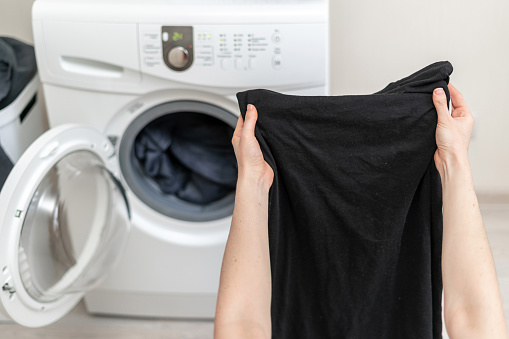 Cropped photo of laundry process. Woman holding black clothes in her hands against white washing machine inside light flat interior