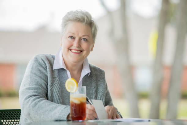 hermosa mujer mayor sonríe mientras escribe carta o tarjeta de felicitación y beber té helado - texas tea fotografías e imágenes de stock