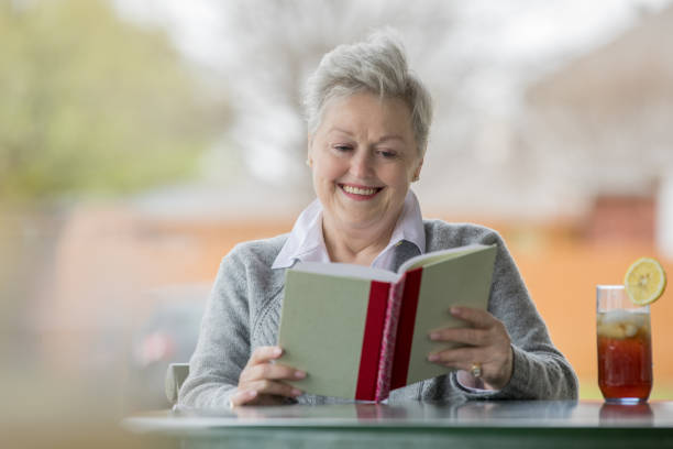 la mujer adulta mayor sonríe alegremente mientras lee un libro y bebe té helado - texas tea fotografías e imágenes de stock