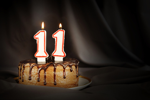 Eleven years anniversary. Birthday chocolate cake with white burning candles in the form of number Eleven. Dark background with black cloth