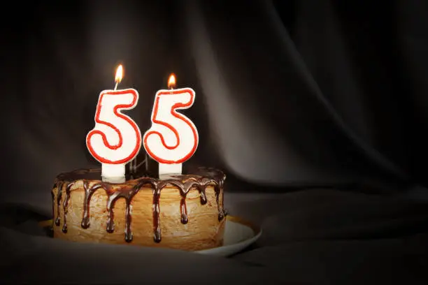 Photo of Fifty five years anniversary. Birthday chocolate cake with white burning candles in the form of number Fifty five. Dark background with black cloth