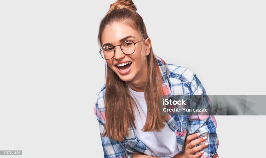 Happy beautiful positive young woman dressed in casual outfit, wearing round transparent eyewear with pleasant broadly smile, looking to the camera and posing over white studio wall. People emotions Adolescence Stock Photo