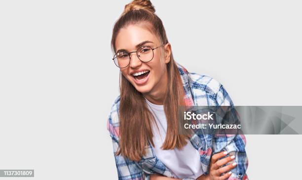 Happy Beautiful Positive Young Woman Dressed In Casual Outfit Wearing Round Transparent Eyewear With Pleasant Broadly Smile Looking To The Camera And Posing Over White Studio Wall People Emotions Stock Photo - Download Image Now