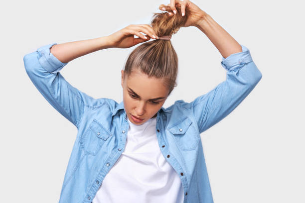 retrato interior de mujer joven recogiendo el pelo en una cola de caballo, con camisa de mezclilla azul y camiseta blanca, posando sobre la pared blanca. adorable mujer rubia hace cola de caballo, anuncia el cabello sano natural - cola de caballo fotografías e imágenes de stock