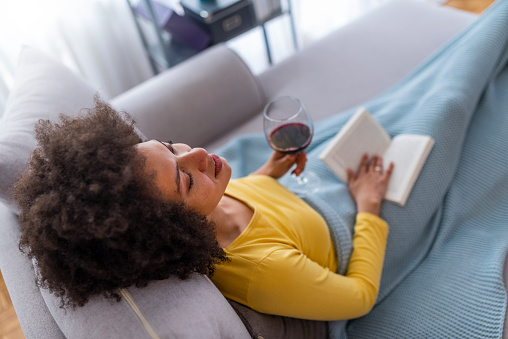 Beautiful young African woman enjoying a glass of wine while relaxing at home. Beautiful young woman reading book near window at home. Woman relaxing in her living room reading book