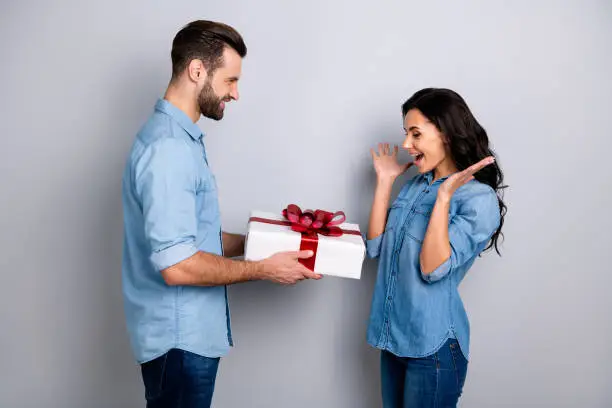 Omg! Profile side view photo of surprised pretty curly-hair lady getting giftbox felicitations from bearded handsome sweetheart waving hands isolated wearing denim clothing on ashy-gray background.