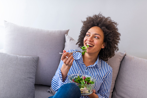 Beautiful woman on the sofa eating a healthy salad. Beautiful healthy woman eating lettuce. Afro American Girl eating organic fresh salad. Close-up of a smiling woman eating a salad in the living-room