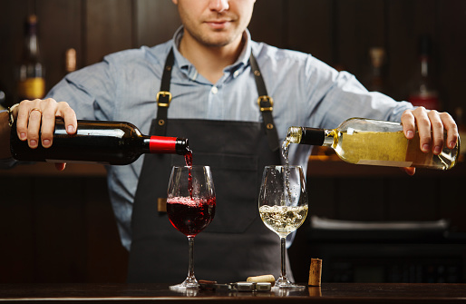 Male sommelier pouring red and white wine into long-stemmed wineglasses