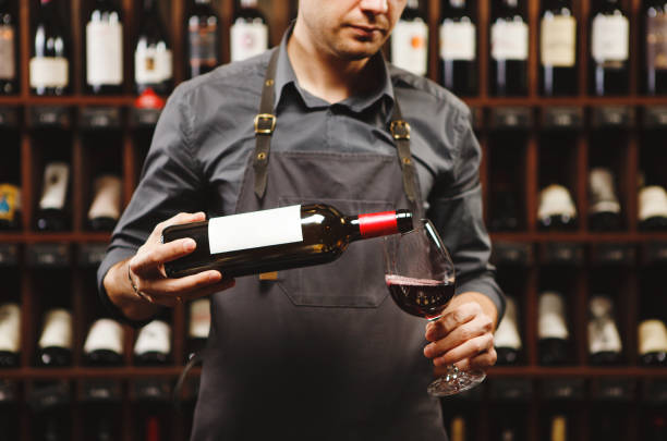 Male sommelier pouring red wine into long-stemmed wineglasses. Male sommelier pouring red wine into long-stemmed wineglasses. Waiter with bottle of alcohol beverage. Bartender on background of shelves with bottles in cellar. sommelier photos stock pictures, royalty-free photos & images