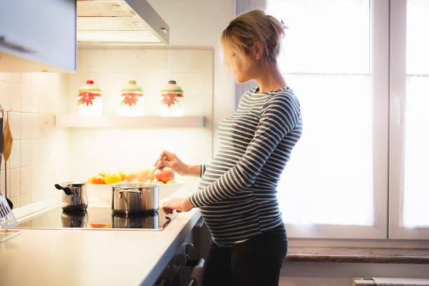 Beautiful Pregnant Woman Cooking Pregnant blond young woman in the kitchen mixing food in pot on electric cooker, with window behind and bowl of fruits. ring tilt stock pictures, royalty-free photos & images