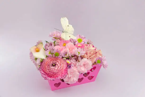 Romantic style floral arrangement in a pink basket with a heart pattern. On top of it a cream-colored butterfly made of feathers. The background light purple.

Meaning of the flowers:

Carnations: Love, Fascination, and Distinction
Geranium: Ingenuity, Melancholy, and True Friendship
Bellis (lawn daisies): Warmth, Care, and Chastity
Buttercup/ranunculum: Humility and neatness
Calla lilly: Resurrection and rebirth, since the plant returns each year after the winter. Faith and purity, especially in the Christian religions