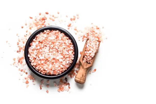 Top view of a black bowl filled Himalayan salt isolated on white background. A wooden serving scoop with salt is beside the bowl and Himalayan salt crystals are scattered on the table. Useful copy space available for text and/or logo. Predominant colors are white and pink. High key DSRL studio photo taken with Canon EOS 5D Mk II and Canon EF 100mm f/2.8L Macro IS USM.