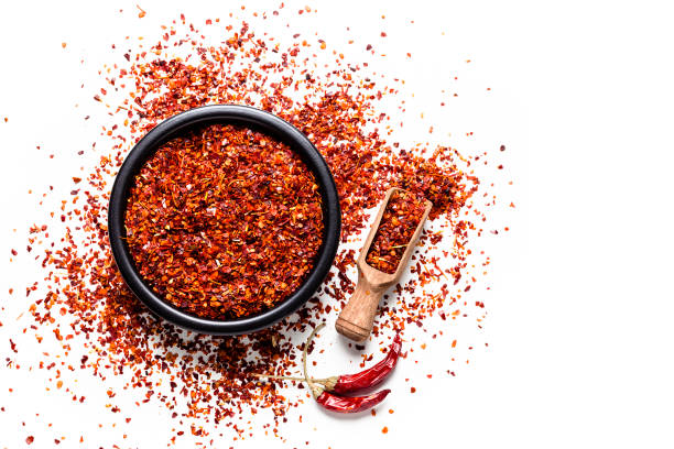 Red chili pepper flakes shot from above on white background Spices: Top view of a black bowl filled with red chili pepper flakes isolated on white background. A wooden serving scoop with pepper flakes is beside the bowl, pepper flakes are scattered on the table. Two dried red chili peppers are beside the bowl. Predominant colors are white and red. High key DSRL studio photo taken with Canon EOS 5D Mk II and Canon EF 100mm f/2.8L Macro IS USM. cayenne powder stock pictures, royalty-free photos & images