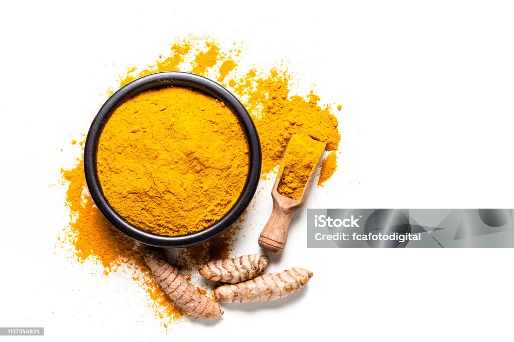 Spices: Turmeric powder and roots shot from above on white background Spices: Top view of a black bowl filled with turmeric powder isolated on white background. A wooden serving scoop with turmeric powder is beside the bowl and turmeric powder is scattered on the table. Fresh organic turmeric roots are beside the spoon. Predominant colors are white and yellow. High key DSRL studio photo taken with Canon EOS 5D Mk II and Canon EF 100mm f/2.8L Macro IS USM. Turmeric Stock Photo