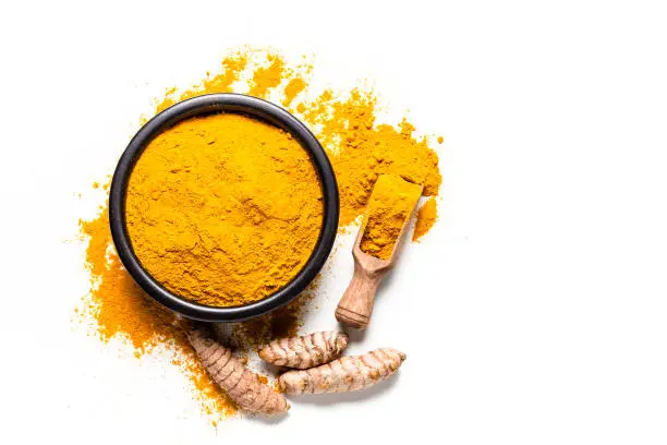 Spices: Top view of a black bowl filled with turmeric powder isolated on white background. A wooden serving scoop with turmeric powder is beside the bowl and turmeric powder is scattered on the table. Fresh organic turmeric roots are beside the spoon. Predominant colors are white and yellow. High key DSRL studio photo taken with Canon EOS 5D Mk II and Canon EF 100mm f/2.8L Macro IS USM.
