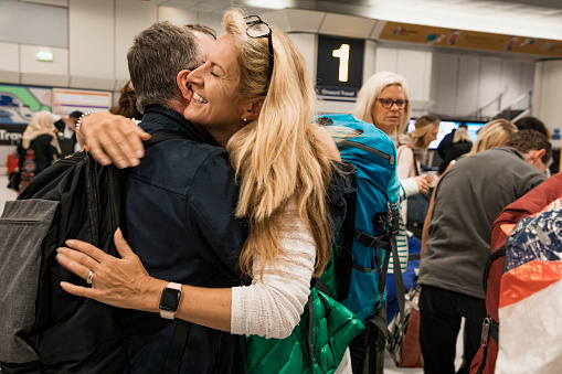 A close-up side-view shot of a man and women hugging eachother at an airport, the woman can be seen smiling with happiness as her friend returns home.