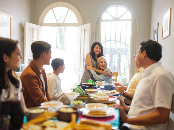 Mexican woman embracing senior woman at lunch table A mexican woman standing and embracing a senior woman sitting at the lunch table with the family, looking happy and smiling. family reunion celebration stock pictures, royalty-free photos & images